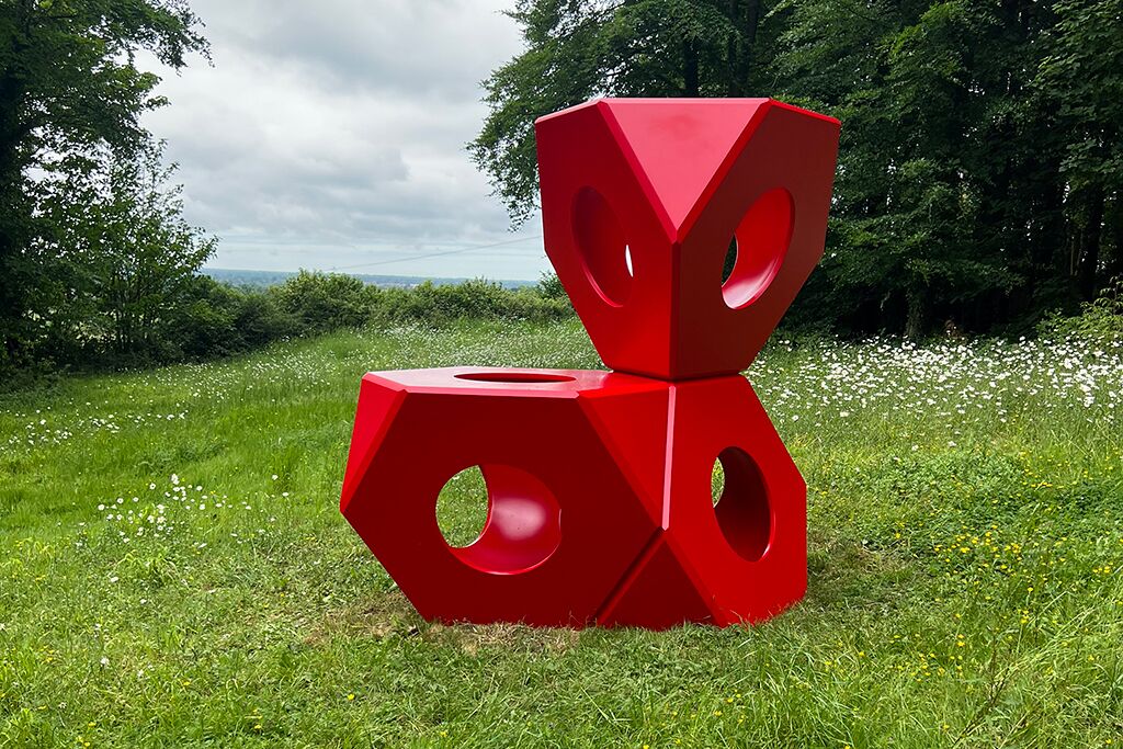 Red geometric sculpture in a grassy field beside trees
